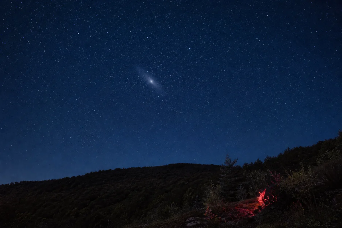 Ciel d&rsquo;automne au-dessus d&rsquo;une crête boisée, la galaxie d&rsquo;Andromède visible comme un fuseau pâle